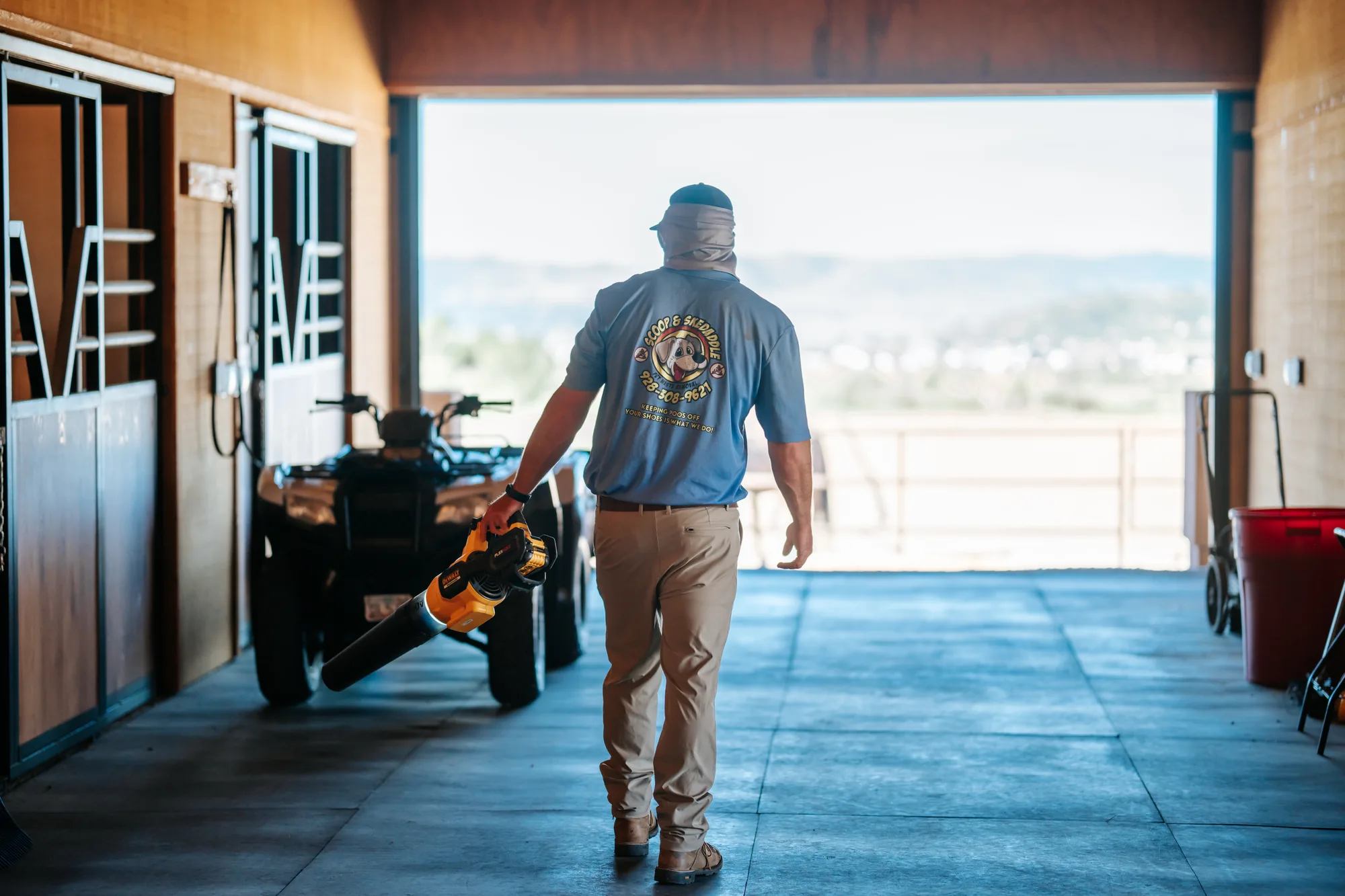 Scoop & Skedaddle team member walking a barn aisle with a blower, backlit by the open doorway