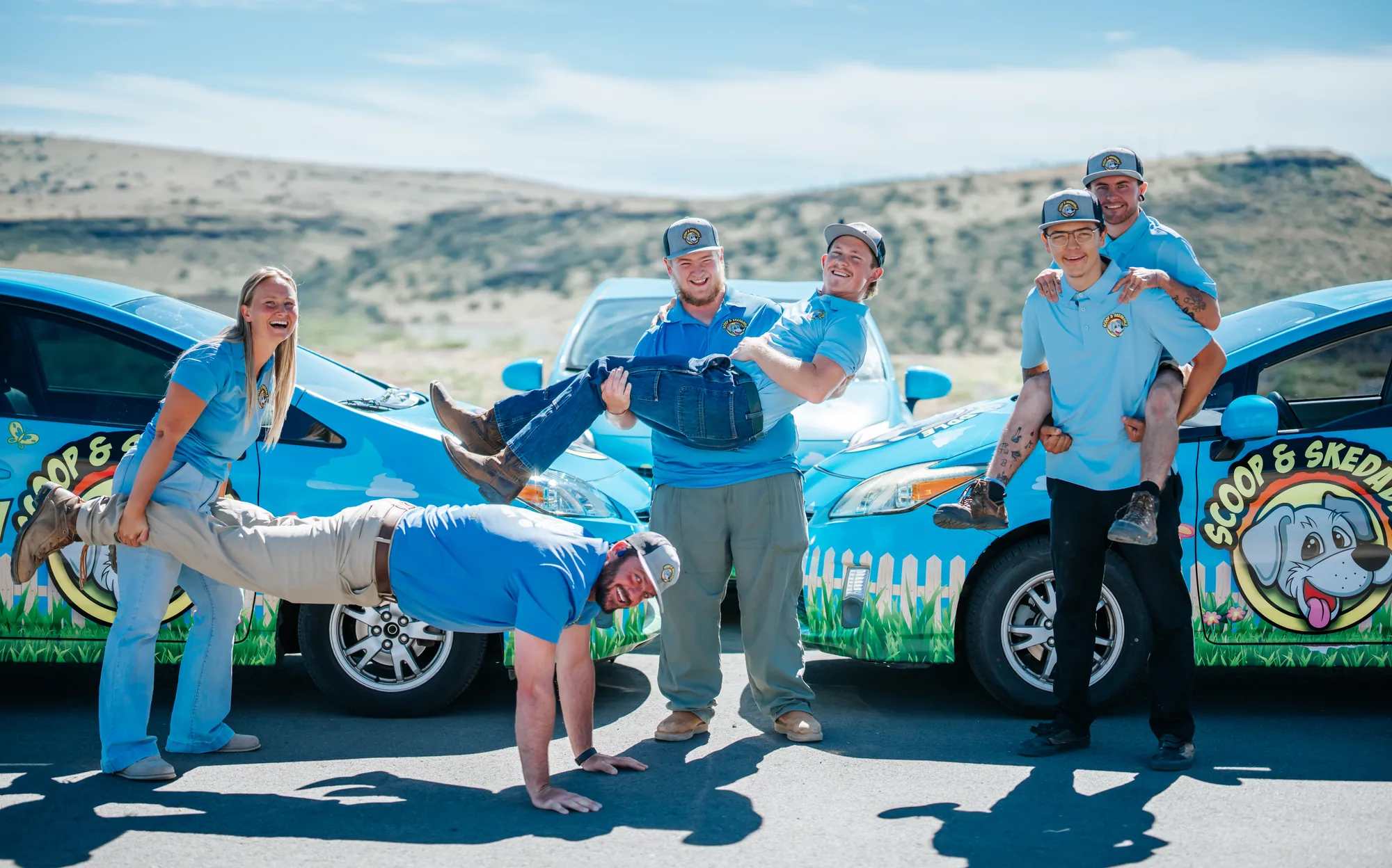 The Scoop & Skedaddle crew clowning around in front of the company cars: a wheelbarrow walk, a bridal carry, and a piggyback