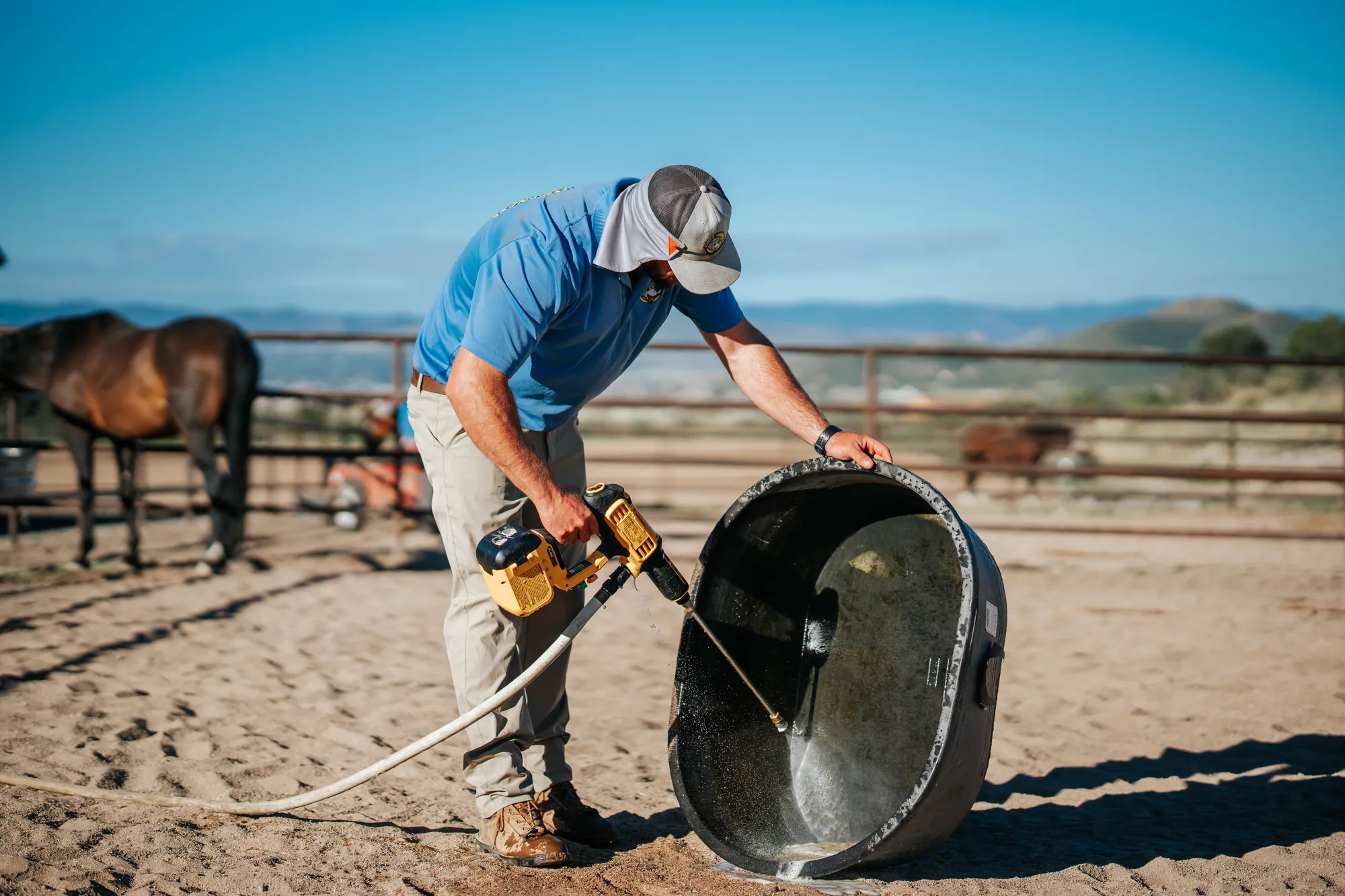 Scoop & Skedaddle team member scrubbing a stock tank with a drill brush, horses in the corral behind