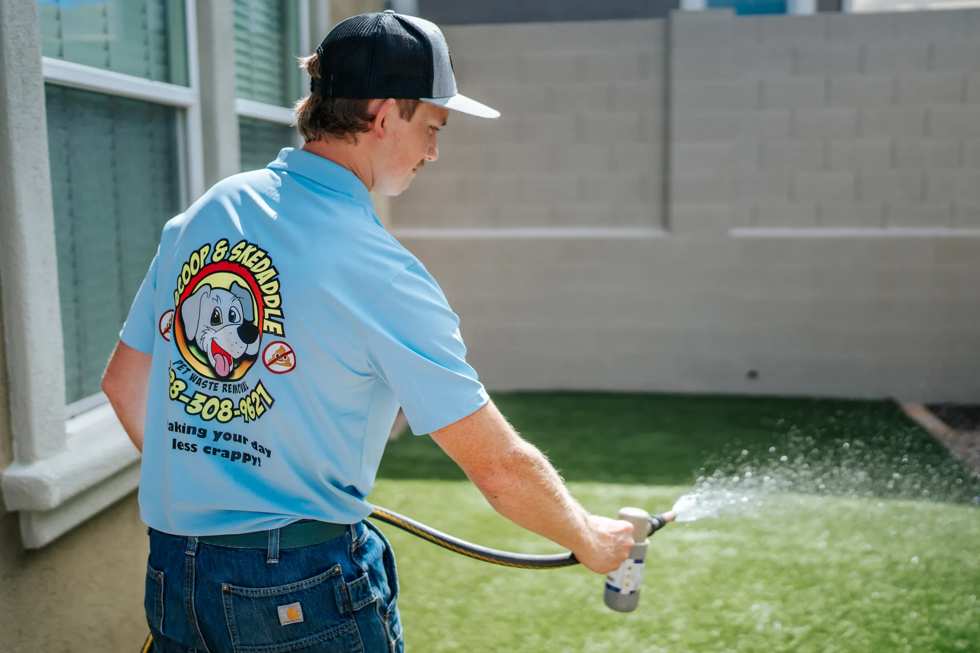 Scoop & Skedaddle technician rinsing artificial turf, the company logo across the back of his shirt