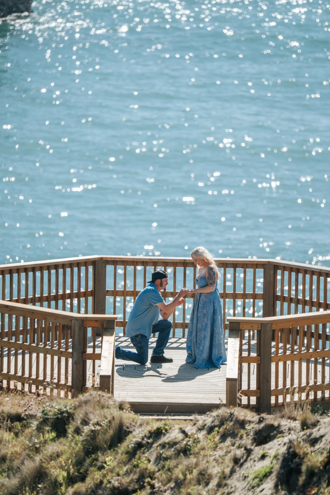 Surprise proposal on a wooden overlook deck above the ocean, he kneels as she says yes