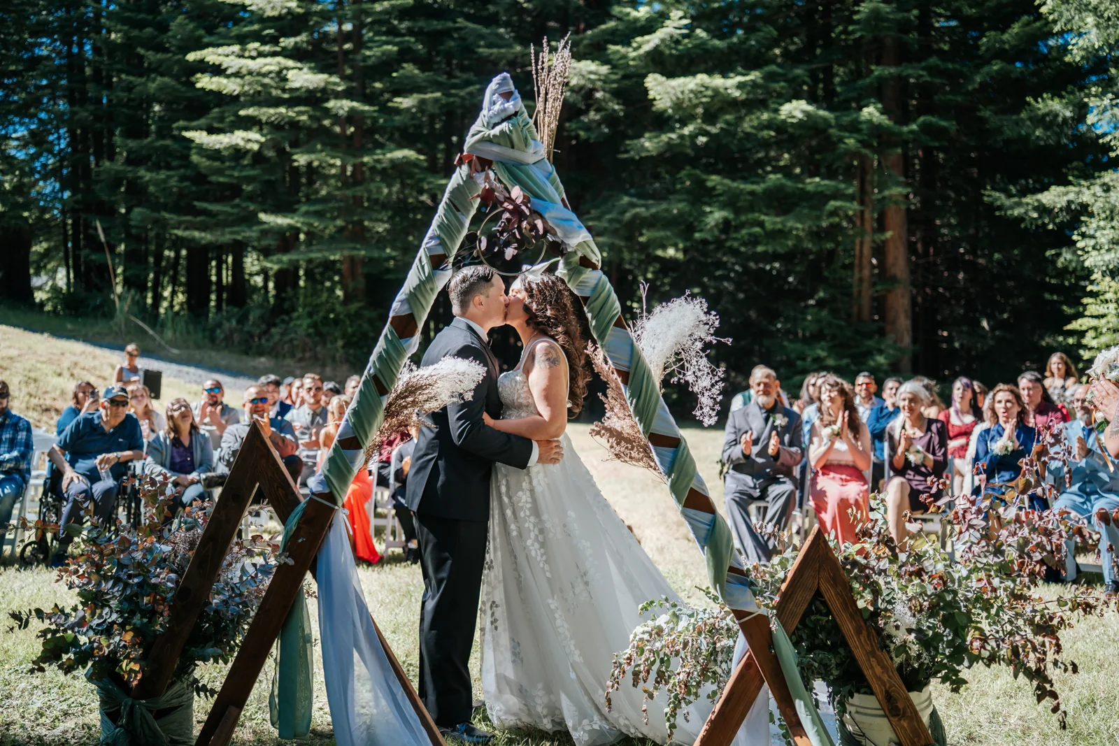 Bride and groom share their first kiss under a wooden triangle arch as guests look on at an outdoor forest ceremony