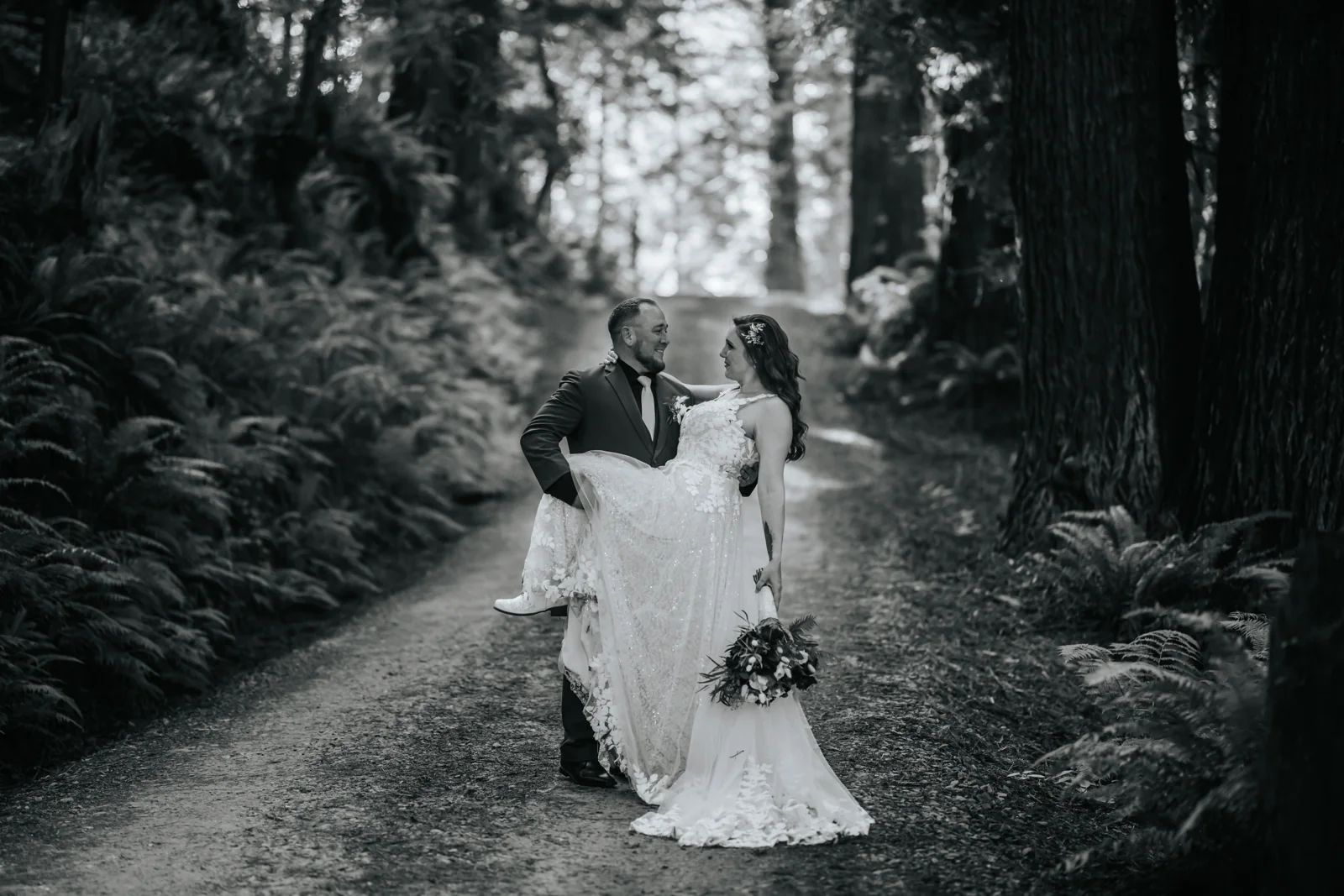 Black and white portrait of a bride and groom walking a fern-lined forest trail