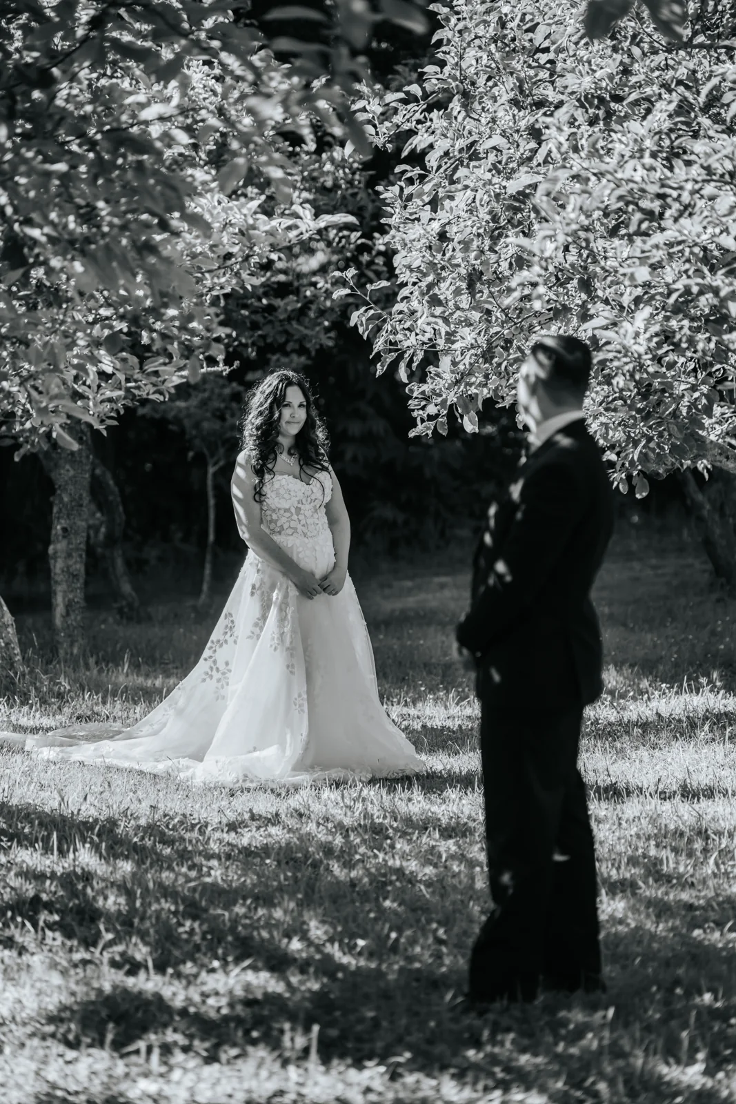 Black and white first look, the bride waiting under a tree as the groom turns to see her