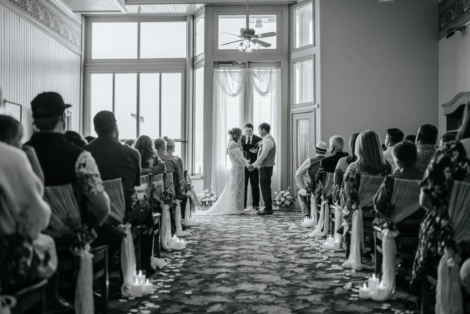 Black and white view down the aisle of an indoor ceremony, the couple framed by tall windows and lit candles