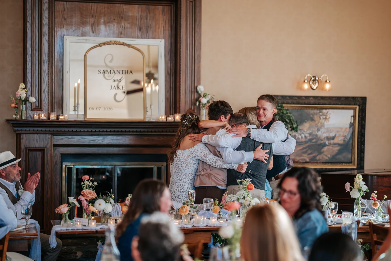 Bride and groom embraced by family in front of a fireplace at their reception